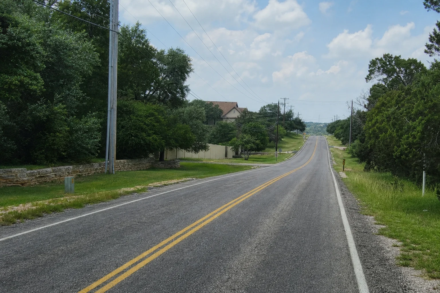 Two-lane road with double yellow lines curving through Hill Country oaks and cedars, past a limestone wall, fence and house, under utility lines and a cloudy sky