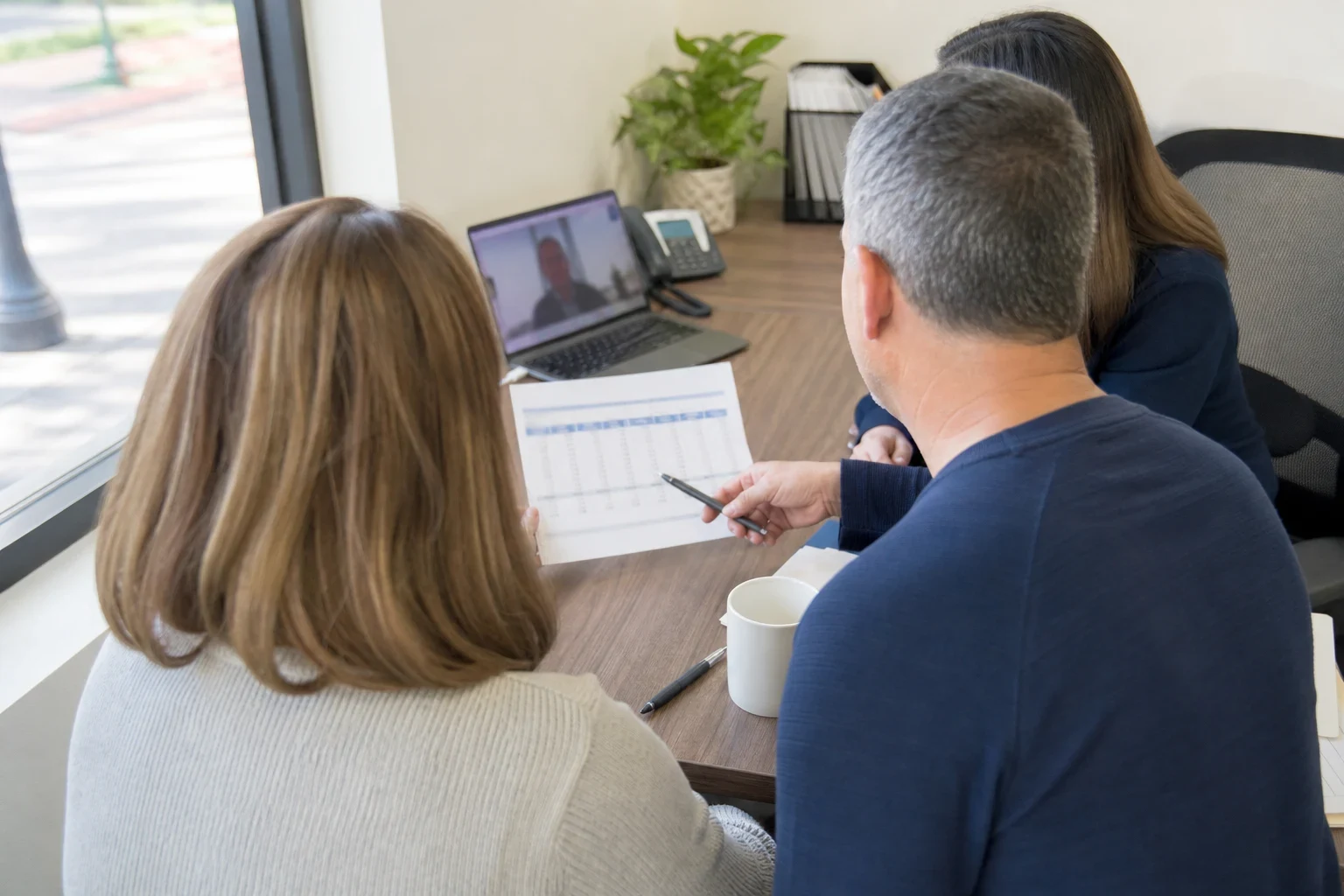 Couple seen from behind at an office desk by a window, reviewing a printed table of figures with a pen, a laptop video call, phone, plant and coffee mug nearby